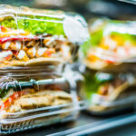 Clear plastic salad containers in a refrigerated deli display, filled with greens, tomatoes and toppings.