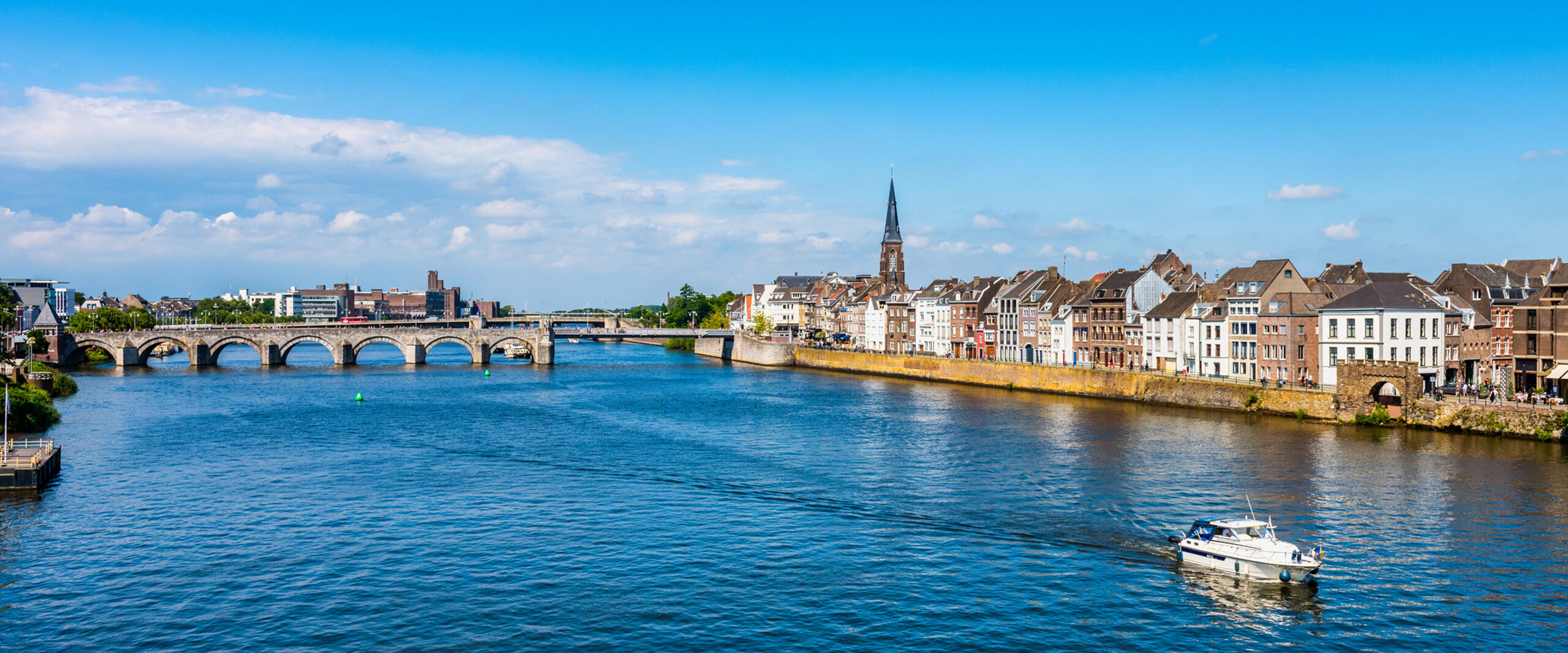 Wide river with a stone-arched bridge and a row of colorful riverside houses under a blue sky.