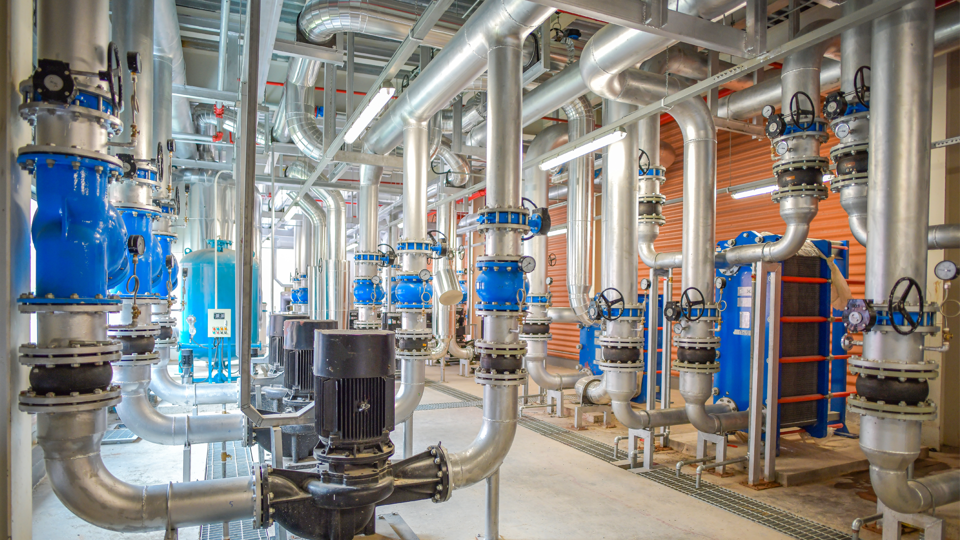 Industrial mechanical room with a network of silver pipes, blue valve assemblies, gauges, and pumps in an industrial processing setup.