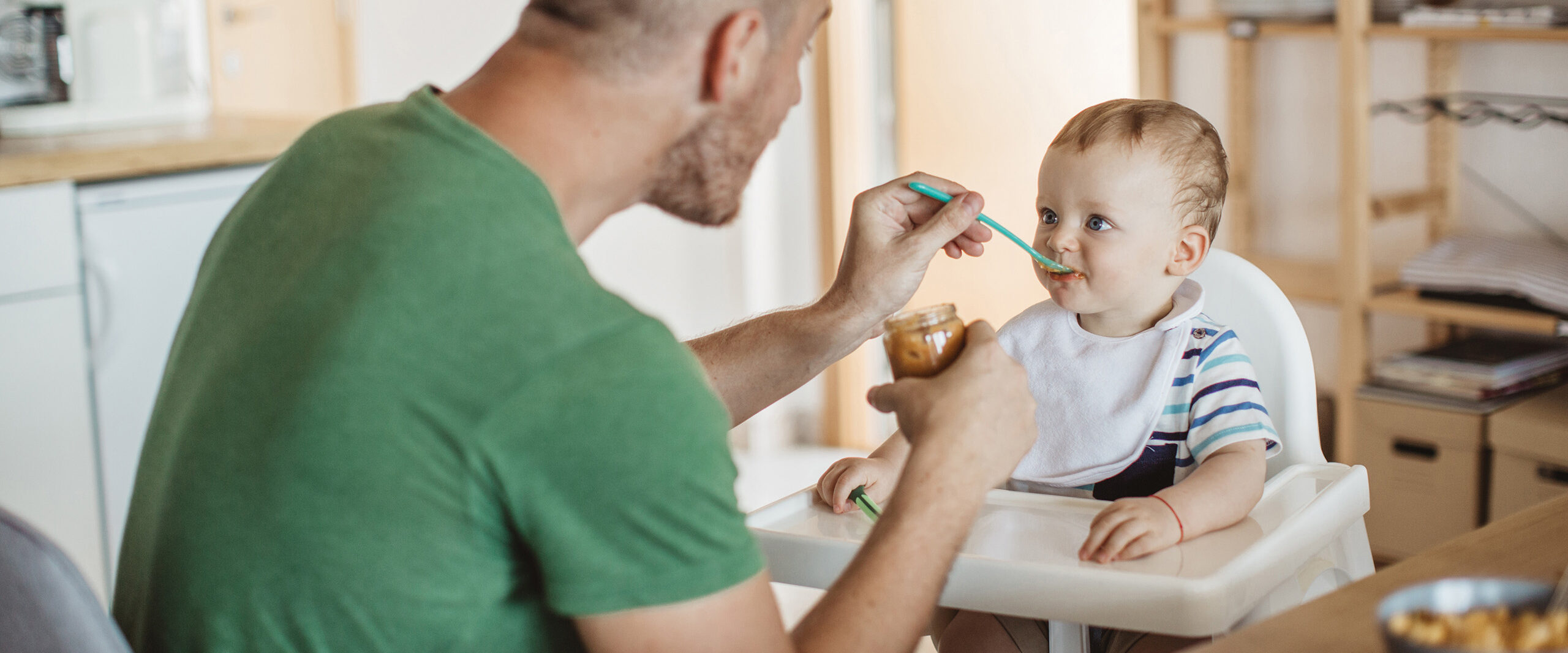 Parent feeding a baby at a high chair with a spoon and jar of baby food in a kitchen.