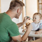Father feeding a baby in a high chair with a spoon and jar of peanut butter at the table in a kitchen.