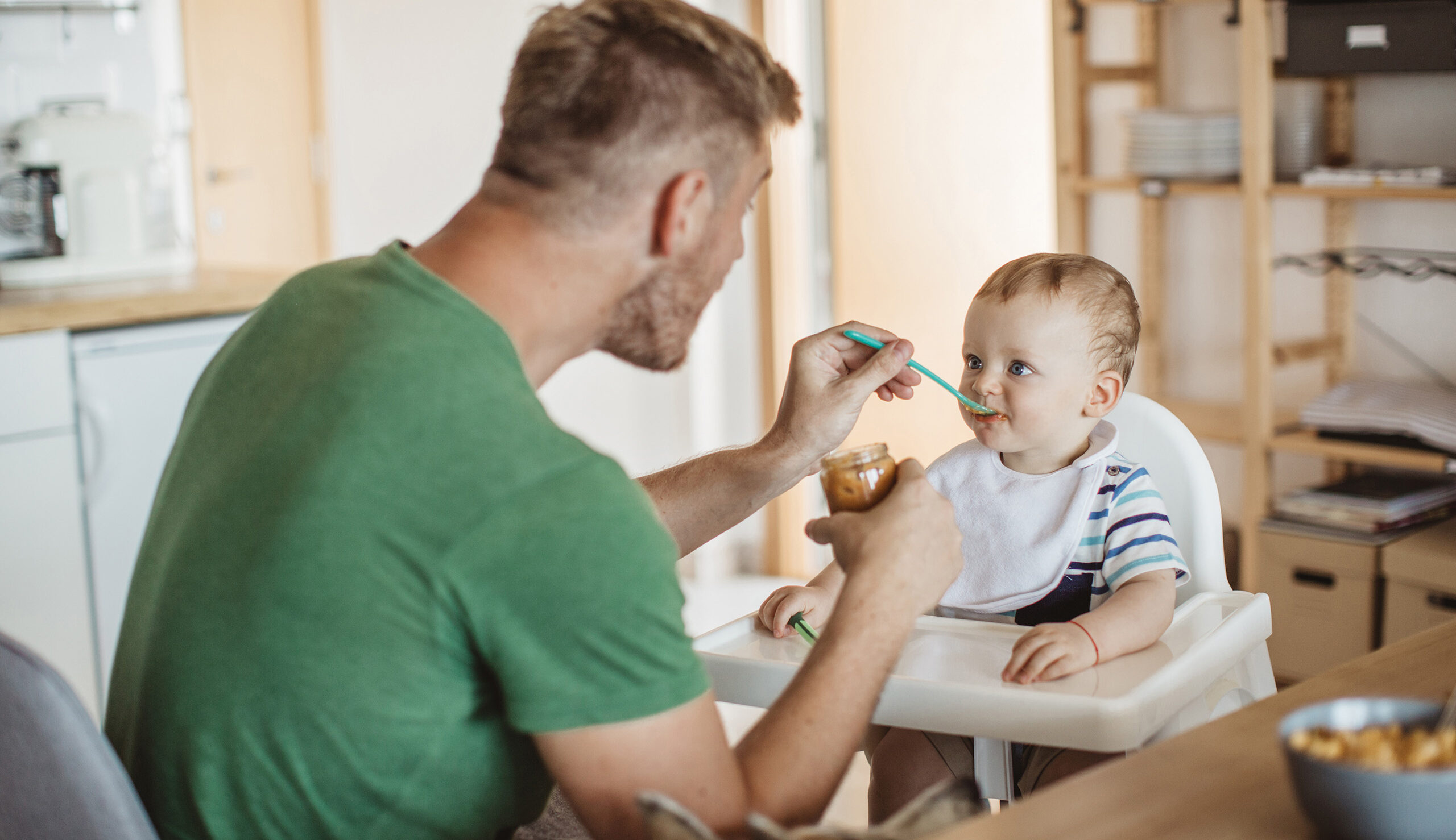 Father feeding a baby in a high chair with a spoon and jar of peanut butter at the table in a kitchen.