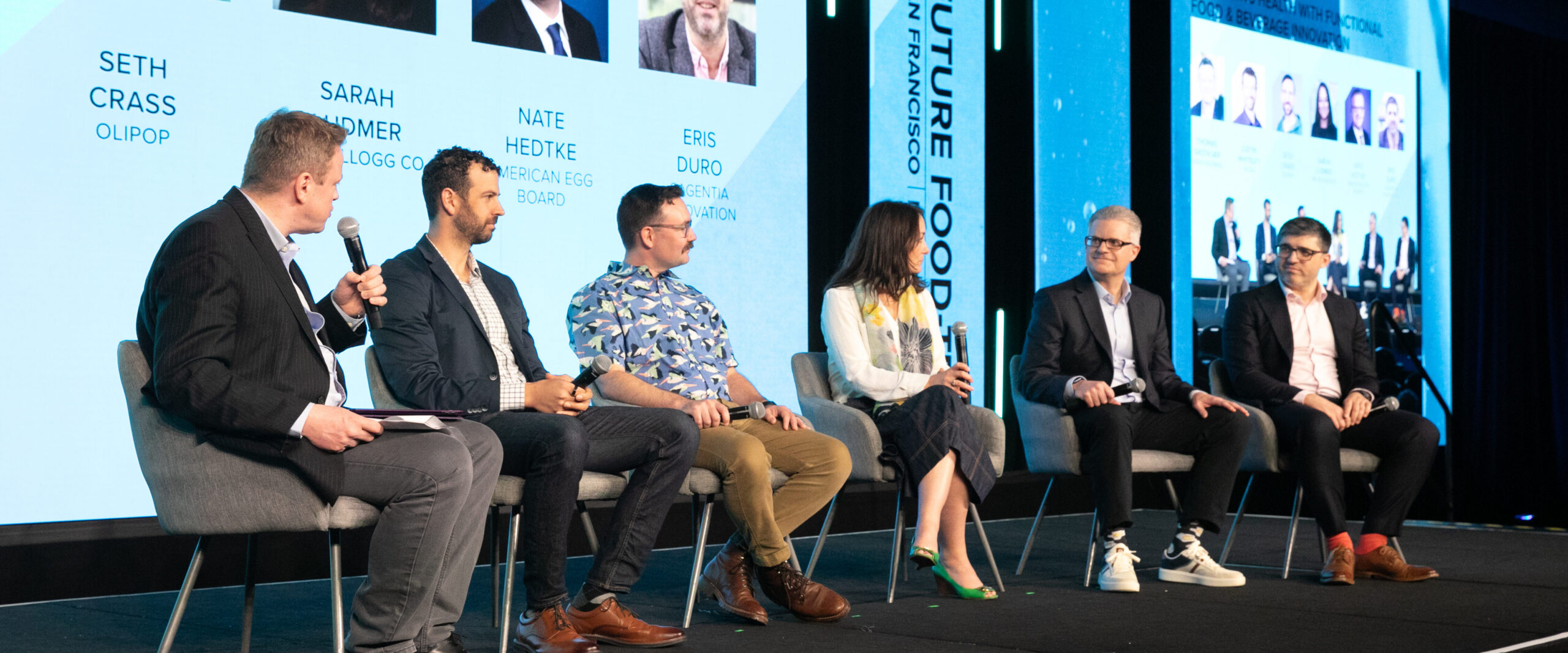Panel discussion with six speakers on stage, each holding a microphone, against a blue backdrop displaying names and images from the event