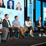 Six-panel discussion on a stage, each person seated in a chair with microphones, blue backdrops and portraits behind them.