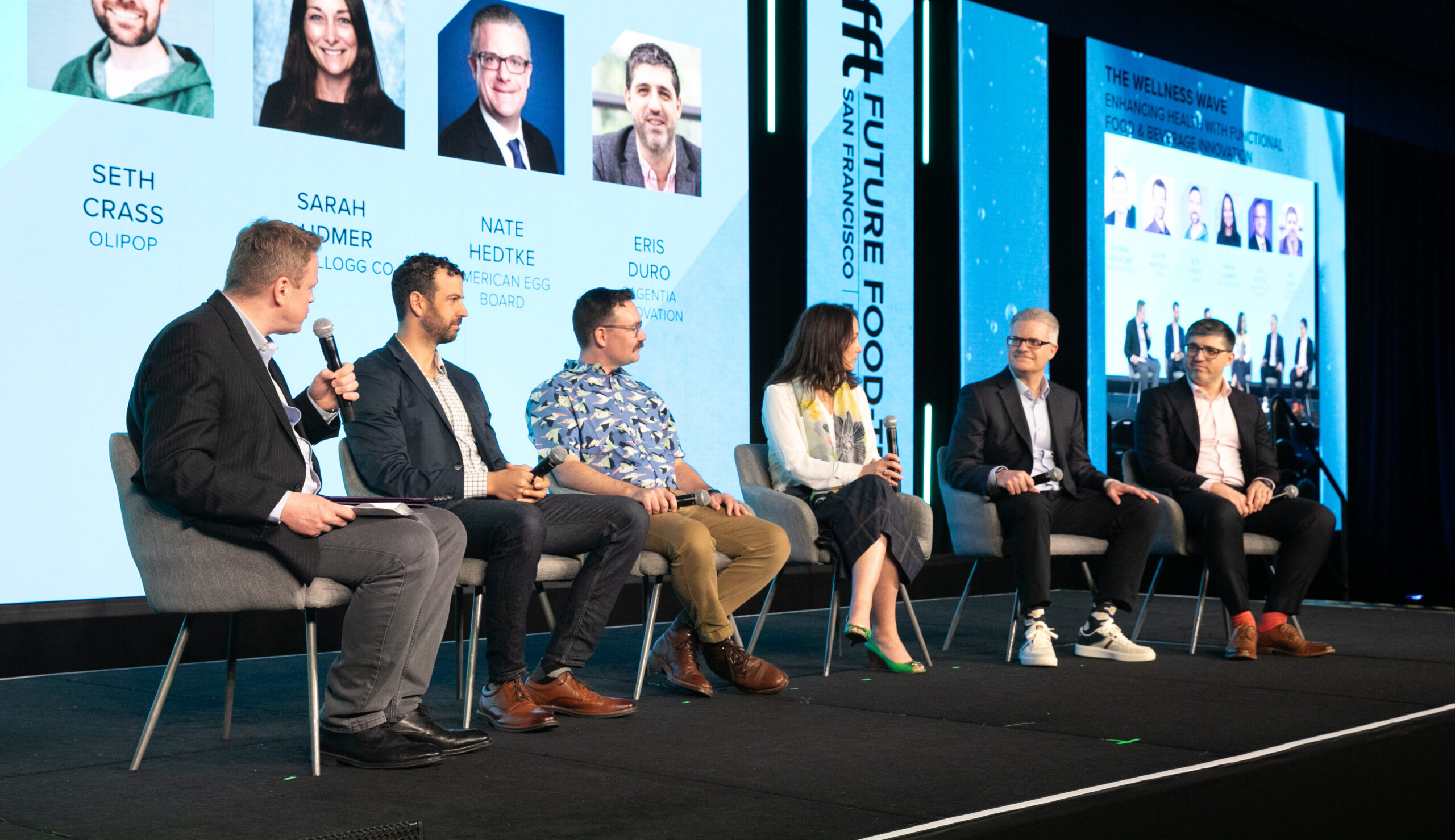 Six-panel discussion on a stage, each person seated in a chair with microphones, blue backdrops and portraits behind them.