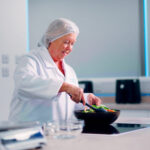 A woman in a white lab coat and hairnet plates a fresh salad in a clean kitchen setting.