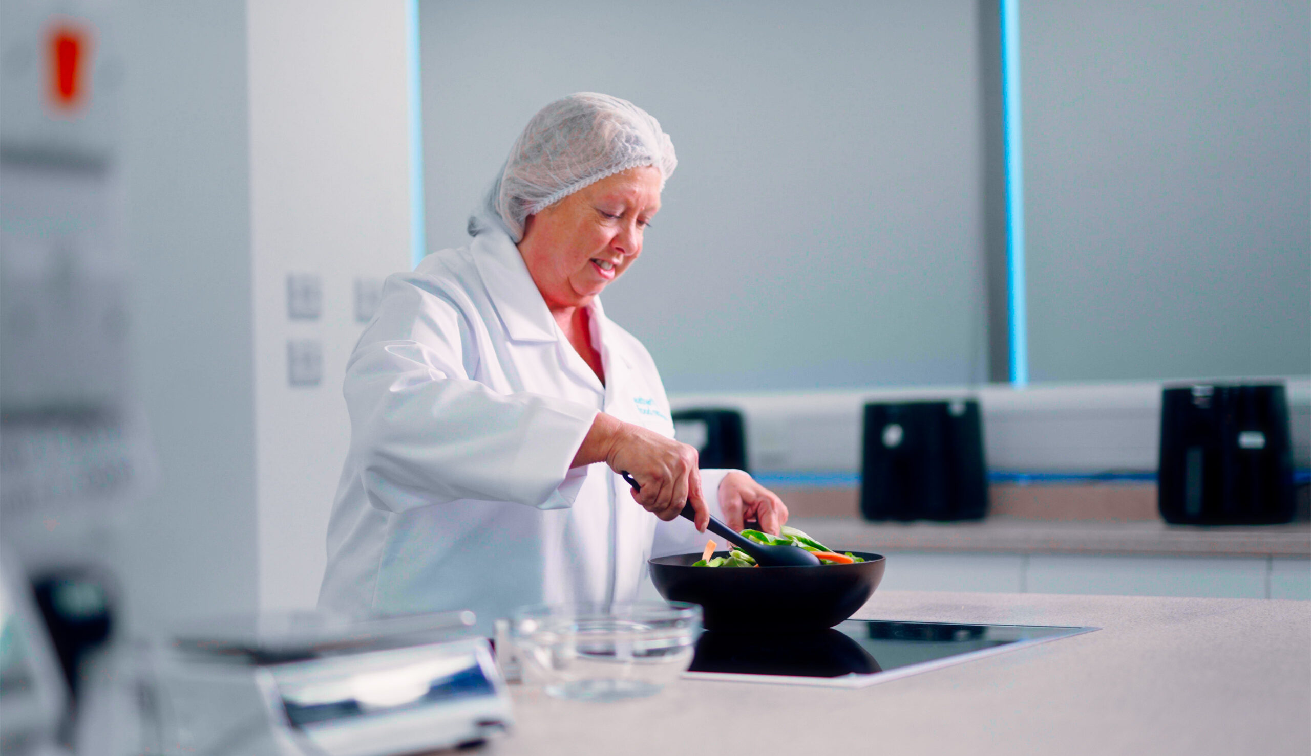 A woman in a white lab coat and hairnet plates a fresh salad in a clean kitchen setting.