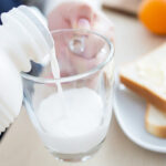 Pouring milk into a glass with buttered toast on a plate in the background, creating a breakfast scene.
