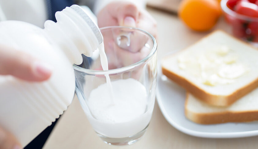 Pouring milk into a glass with buttered toast on a plate in the background, creating a breakfast scene.