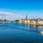 Riverside town with a stone arch bridge across a calm blue river under a bright sky.