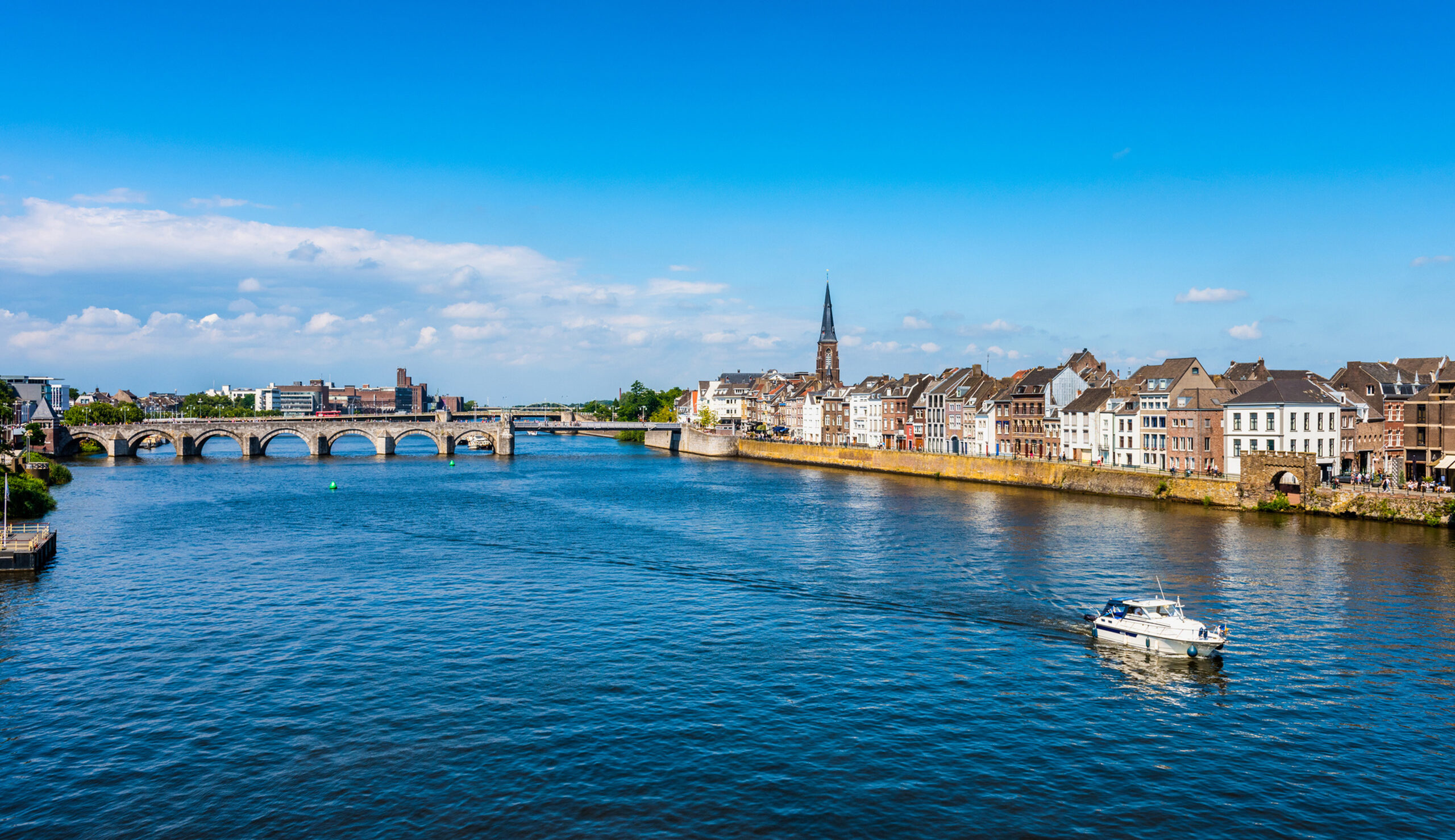 Riverside town with a stone arch bridge across a calm blue river under a bright sky.