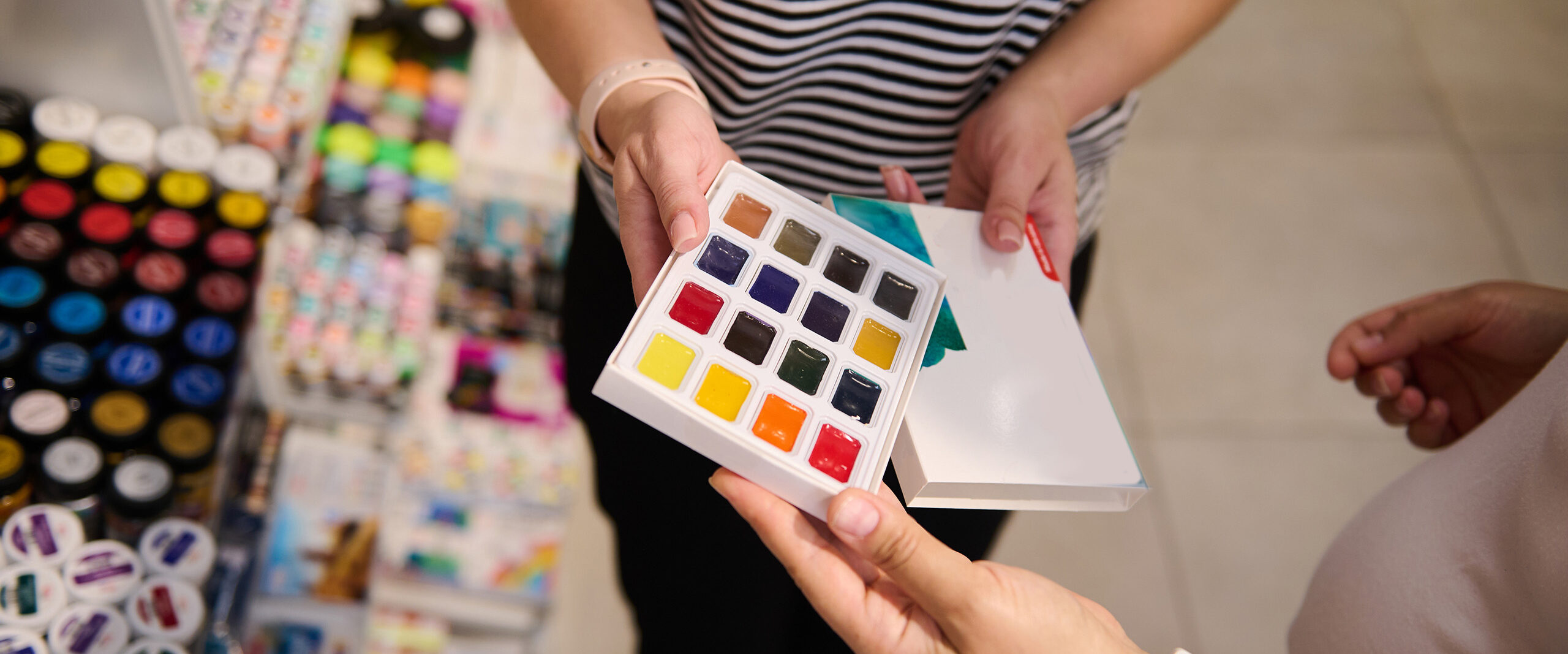 Person holds a square swatch book with 24 colorful paint blocks in a craft store, shelves in the background
