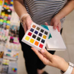 Person holding a color swatch palette with 24 paint squares in a craft store background blur of supplies.