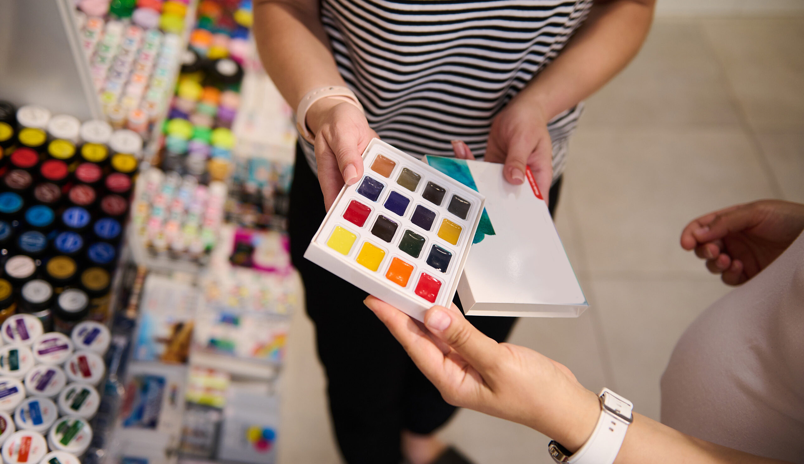 Person holding a color swatch palette with 24 paint squares in a craft store background blur of supplies.