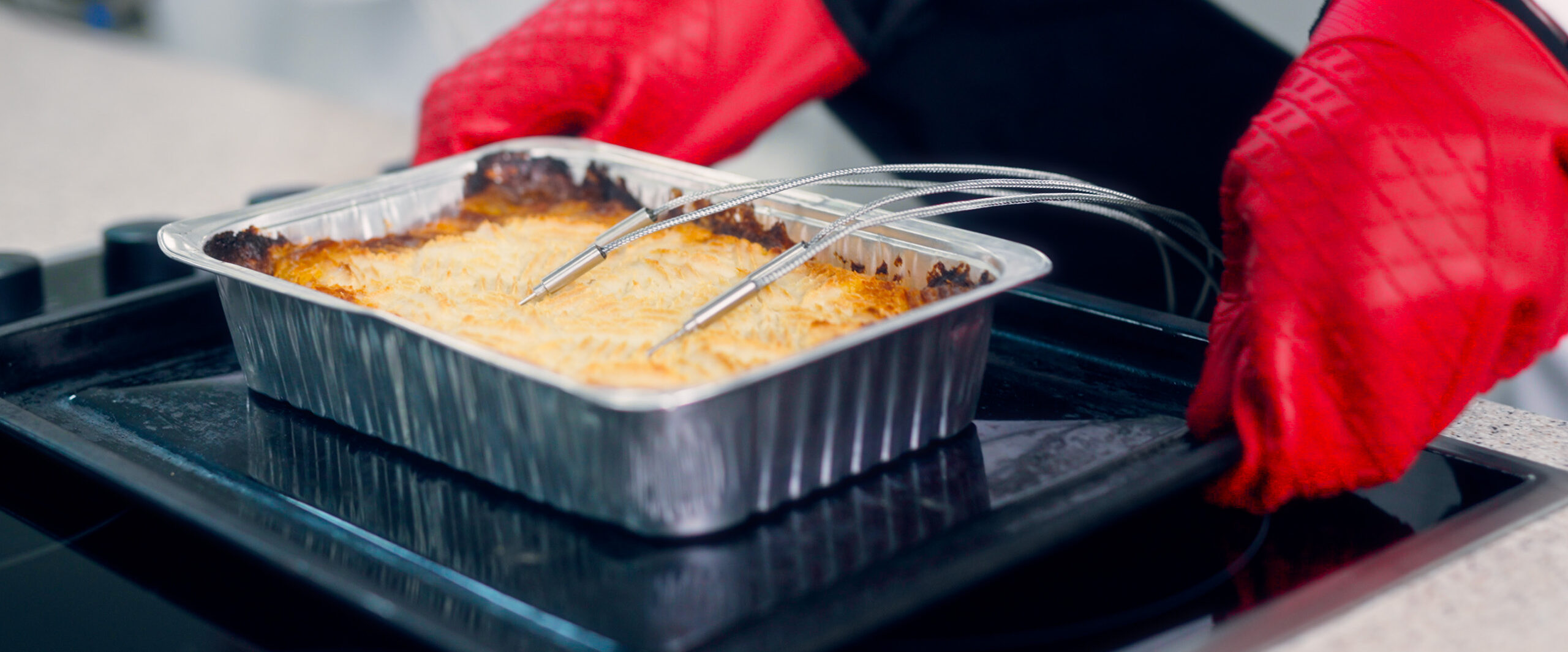 Aluminum baking pan on a stovetop with a golden baked crust and two metal skewers resting on top, red oven mitts nearby