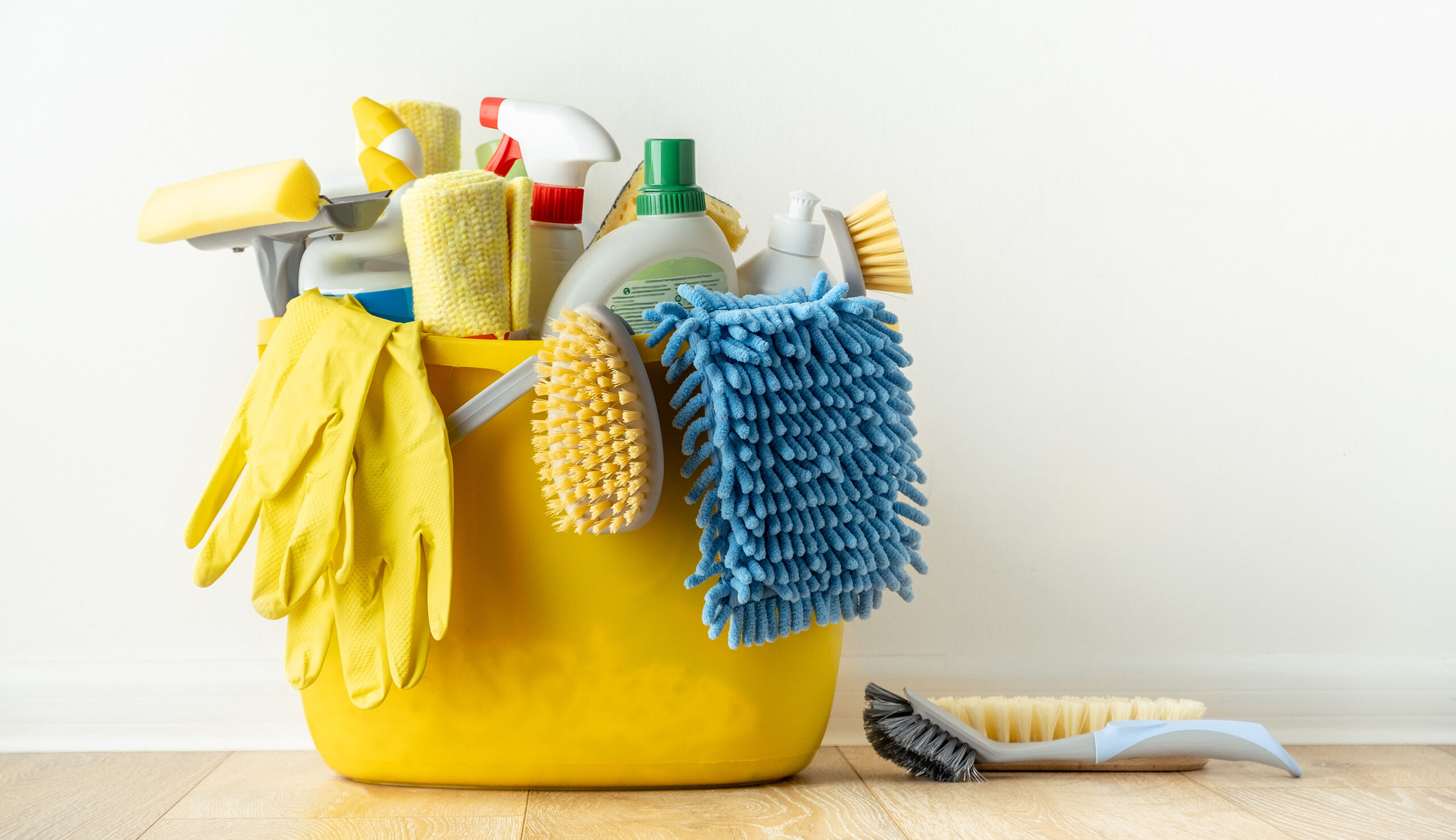 Yellow bucket overflowing with cleaning supplies: gloves, brushes, sponges, and a spray bottle in view.