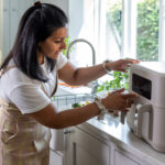 Woman loading or adjusting a bread machine on a kitchen counter by a window, wearing a plaid apron and watch.