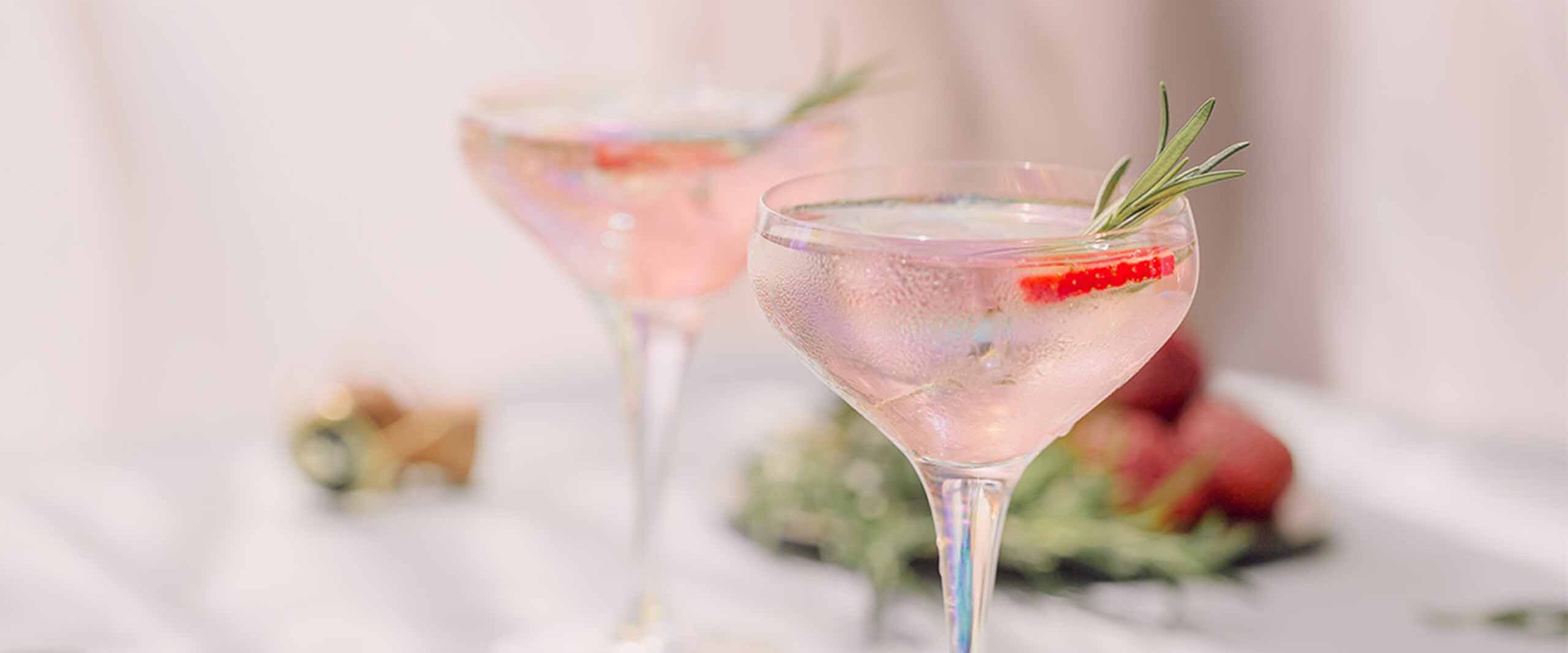 Two pink cocktails in coupe glasses on a bright table, garnished with rosemary sprigs and a strawberry slice Reacting with condensation on the glass.