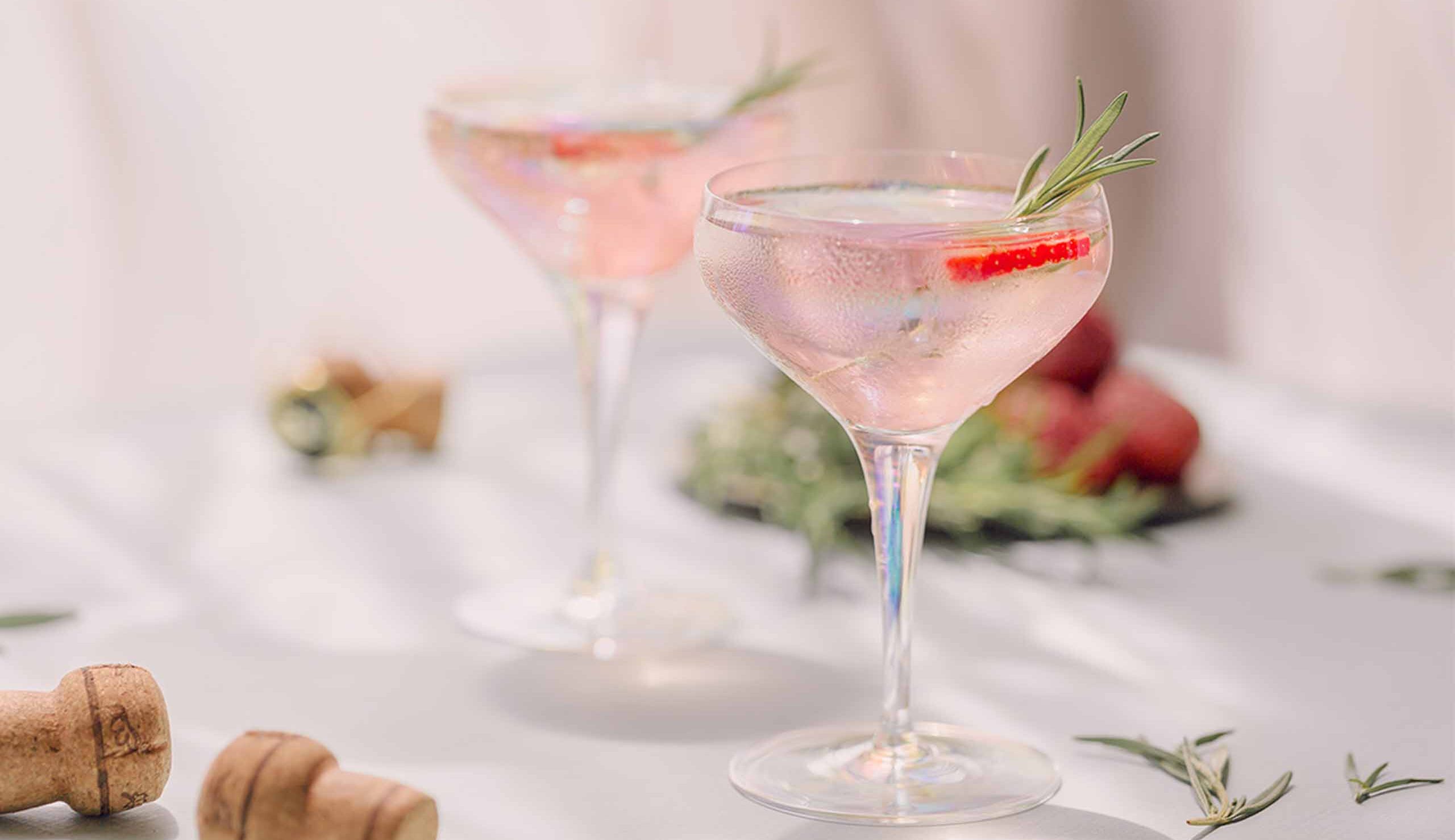 Two elegant cocktail glasses on a white table, garnished with rosemary sprigs and a strawberry slice, in a festive setting.
