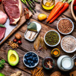 Assorted groceries on a rustic wooden table: raw steak and chicken, tomatoes, carrots, avocado, cheese, grains and legumes in bowls, olive oil, herbs, berries, and a mix of vegetables.