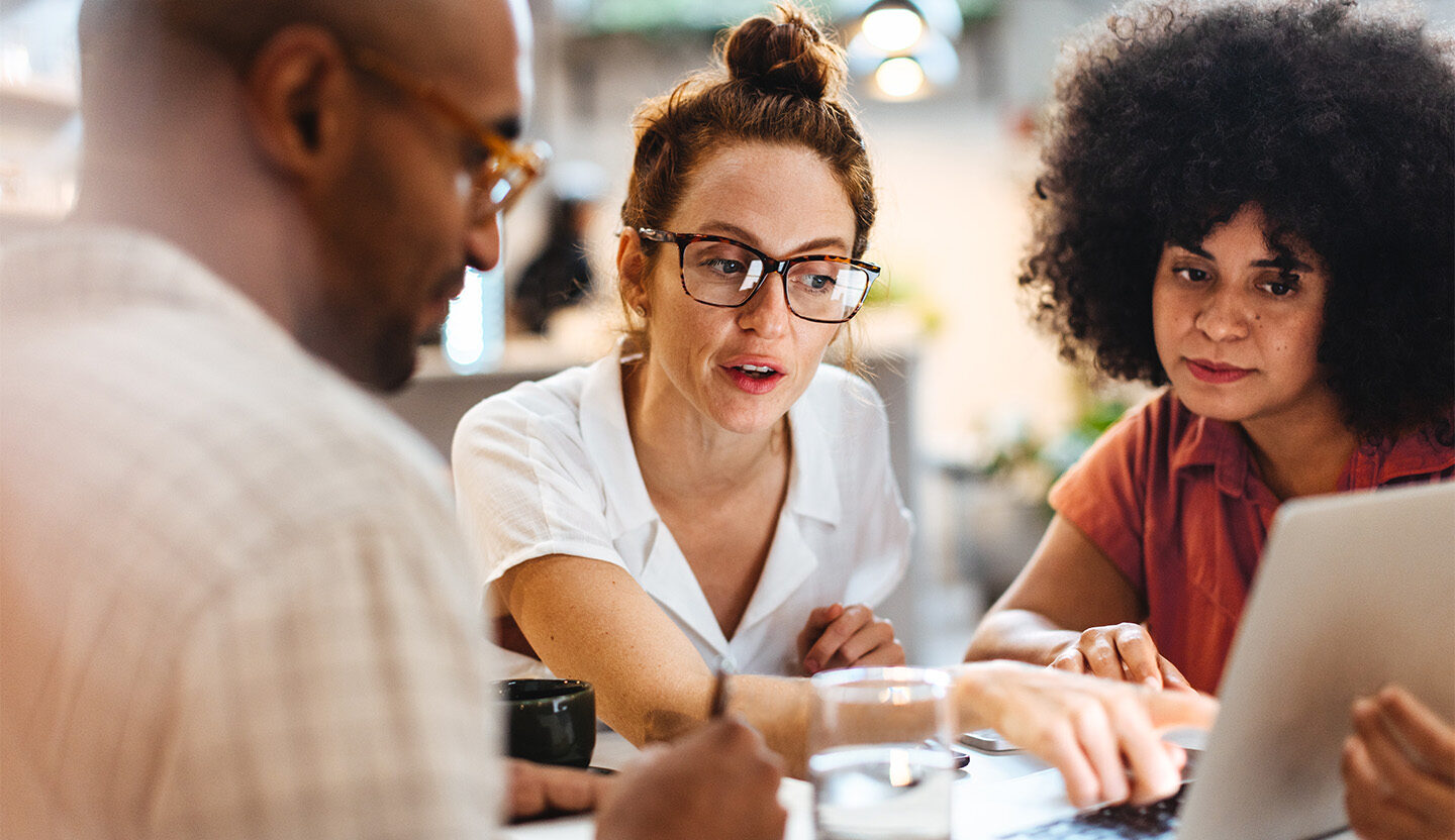 Three coworkers gathered around a table, collaborating with a laptop and notebook.