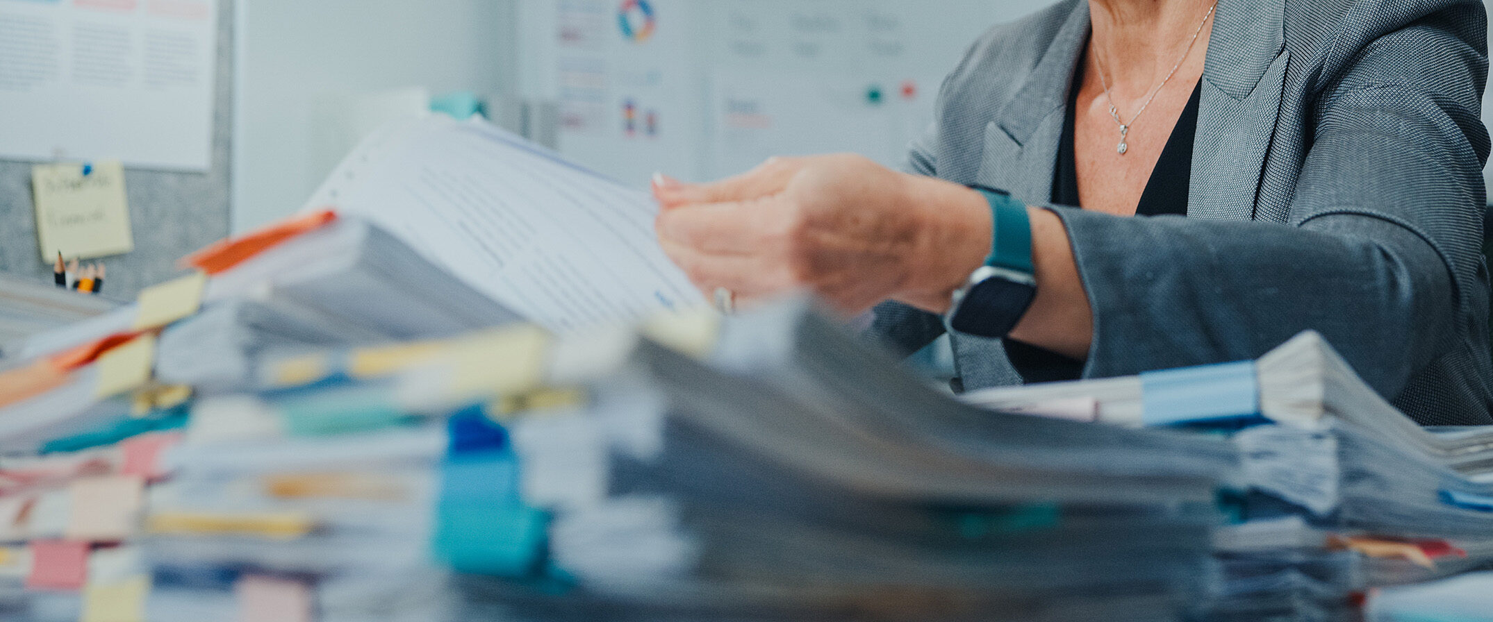 Person in a gray blazer sorts through a large stack of papers and sticky notes at a cluttered office desk.