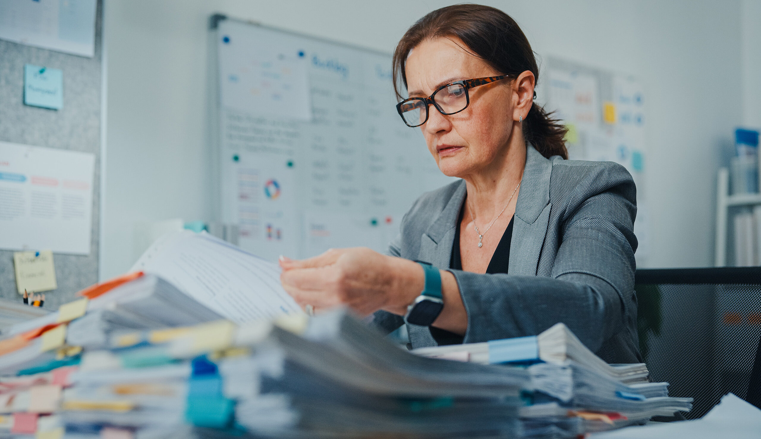 Professional woman with glasses at a cluttered desk, reviewing documents and sticky notes in an office.
