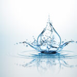 High-speed shot of a blue water splash forming a crown shape with droplets in mid-air against a pale blue gradient background, reflecting on the water surface.