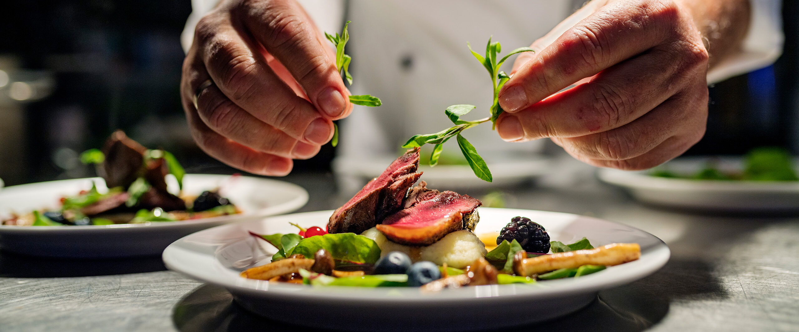 Chef’s hands garnish a plated meat dish with fresh herbs, adding a bright green sprig over slices of meat and vegetables.