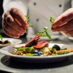 Chef garnishing a plated gourmet dish with fresh greens over slices of meat and berries at a restaurant kitchen.