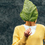 Person in a yellow sweater holds a large green leaf over their face, obscuring their identity against a dark background.