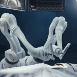 Robotic surgical arms operating on a patient in an illuminated operating room with monitors overhead.