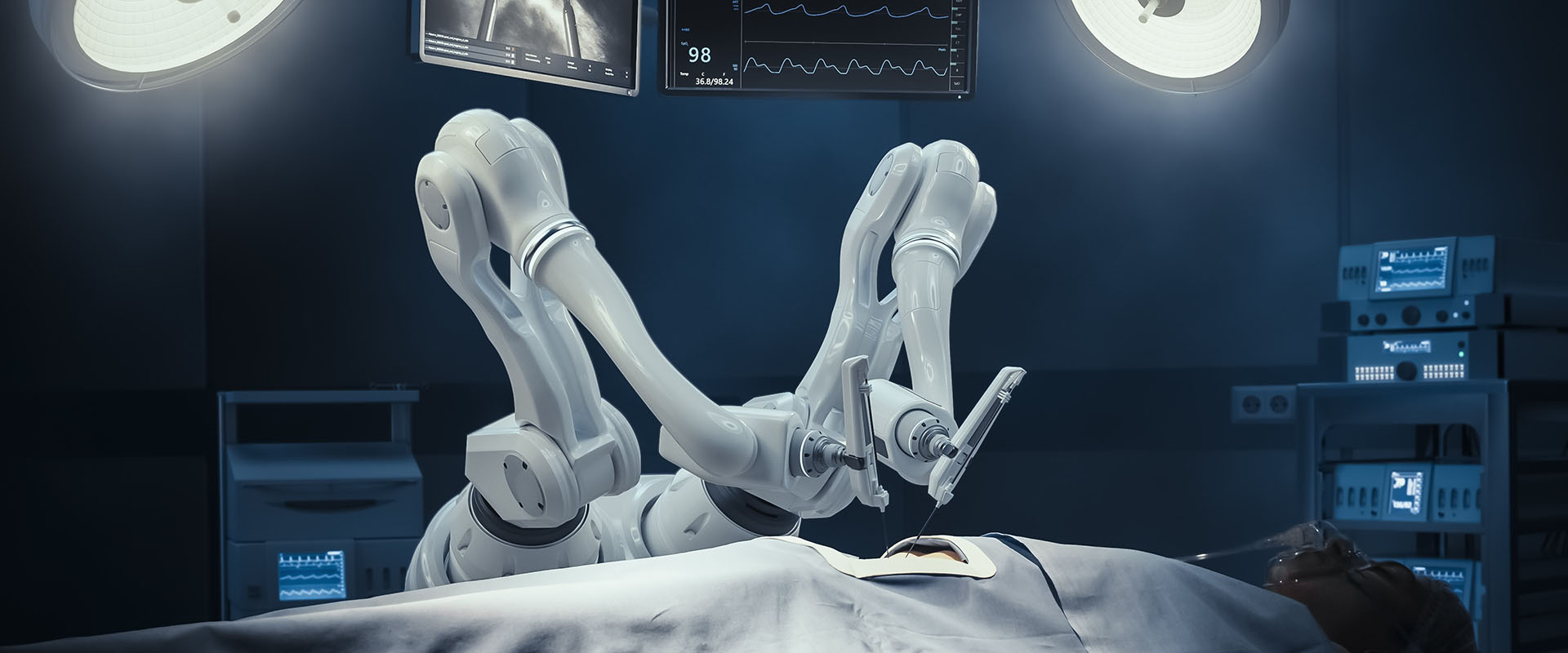 Robotic surgical arms operate over a patient on an operating table under bright overhead lights, with monitors above showing vital signs.