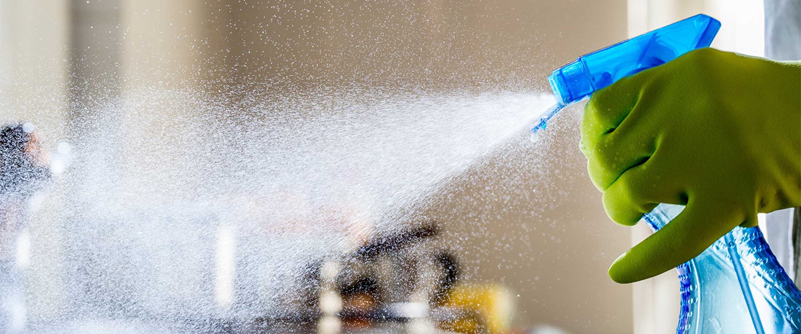 Gloved hand in a bright green glove sprays a blue spray bottle, releasing a fine disinfectant mist across a surface.