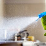 Person wearing a green glove sprays a blue cleaning bottle, releasing a fine cleaning mist toward a surface inside a room.