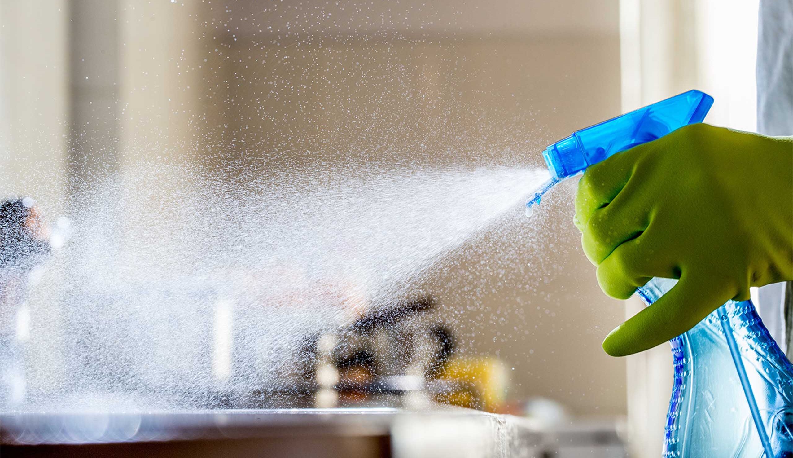 Person wearing a green glove sprays a blue cleaning bottle, releasing a fine cleaning mist toward a surface inside a room.
