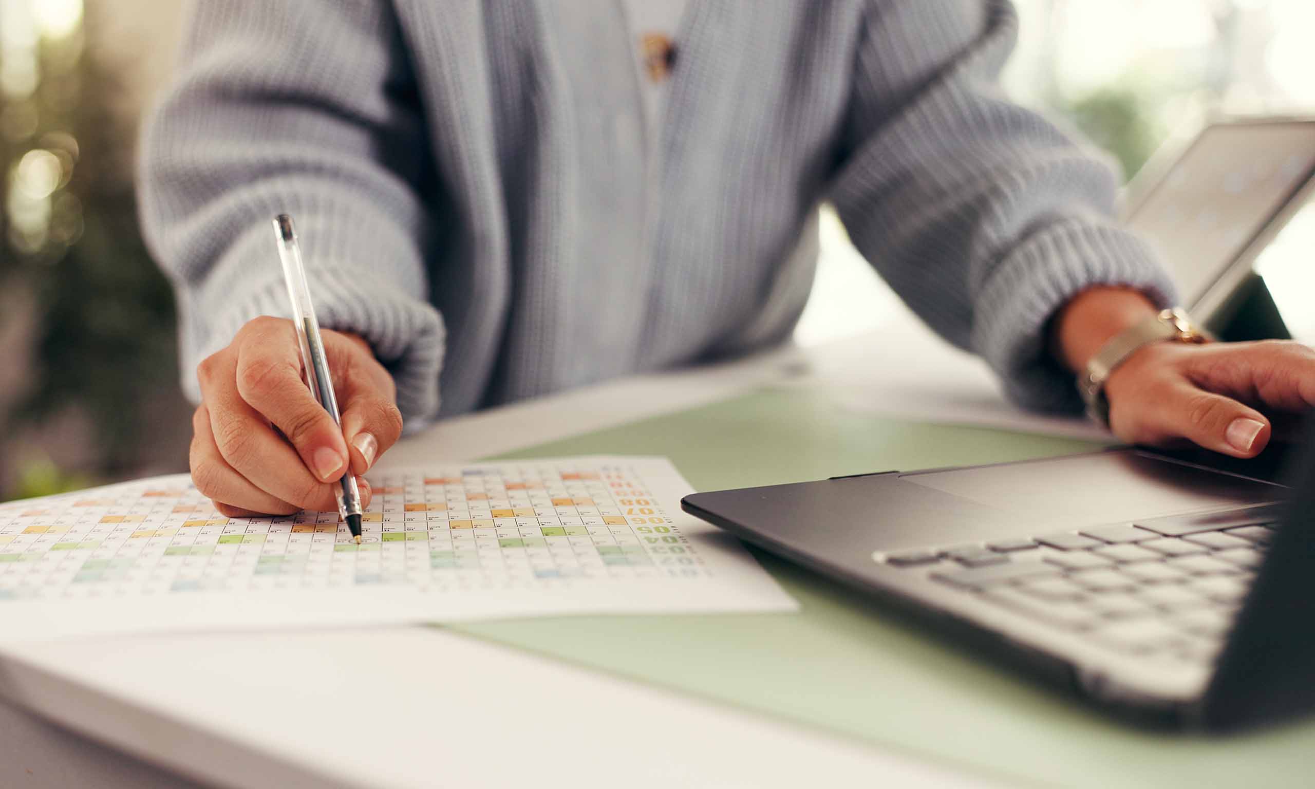 Close-up of a hand with a pen marking a colorful grid on paper at a desk, with a laptop visible to the right.