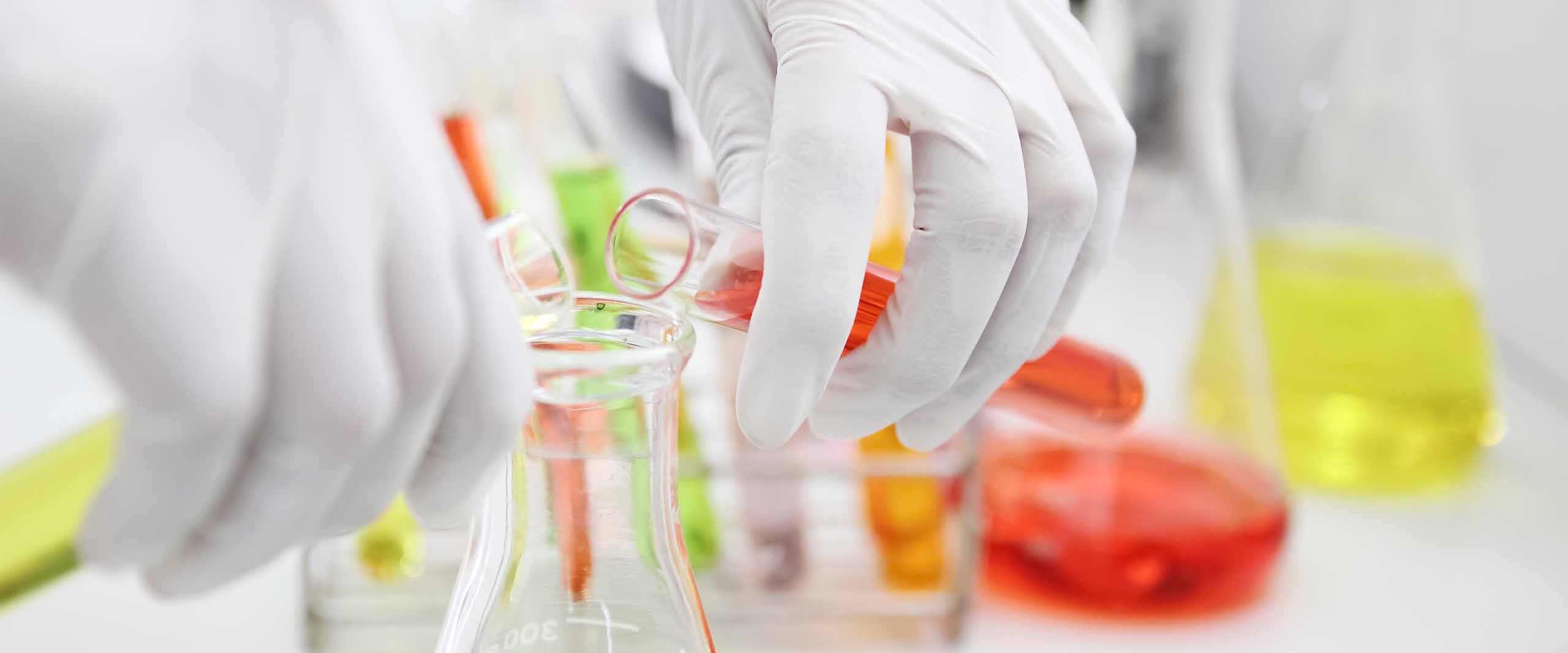 Gloved hand pouring orange liquid from a pipette into a beaker in a bright laboratory setting