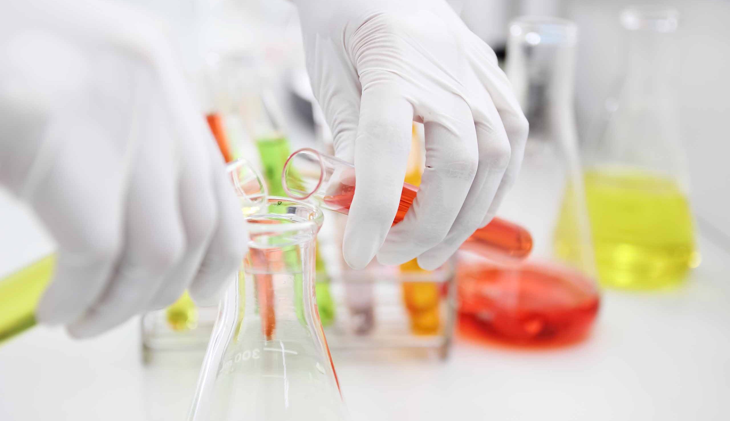 Gloved hand handling a glass Erlenmeyer with orange liquid in a laboratory.