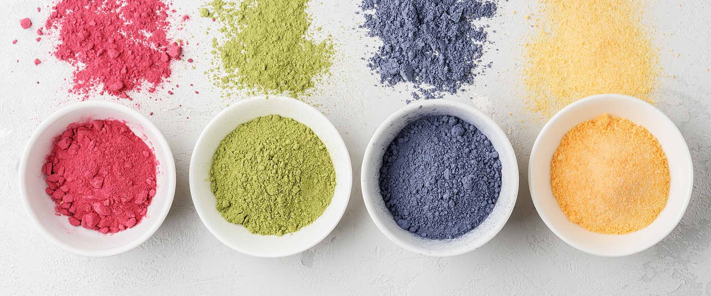 Four bowls contain vibrant pigment powders—pink, green, blue, and orange—on a light surface with scattered powder nearby.