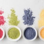 Assorted colored pigment powders spilled from bowls onto a light surface, pink, green, blue, and yellow hues visible at the top and in bowls at the bottom.