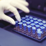 Gloved hand picking up a small blue-lidded test tube from a rack in a lab.