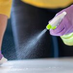 Person wearing purple gloves spraying cleaning solution onto a surface while wiping with a cloth/newspaper for cleaning