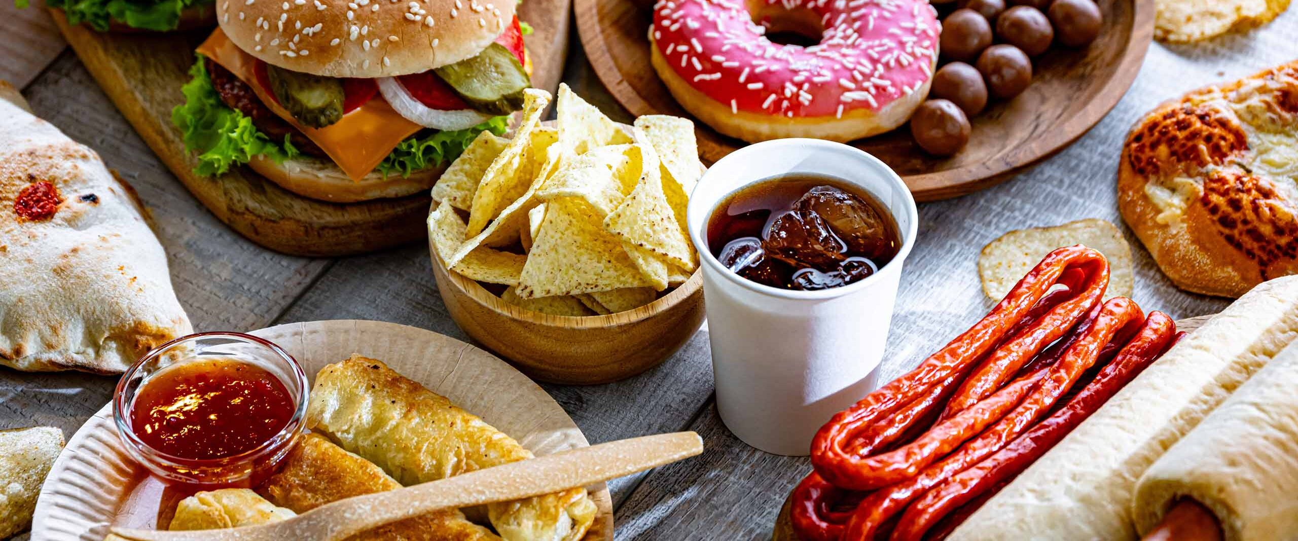 Assorted snack platter on a wooden table: burgers, donuts, tortilla chips, salsa, a cup of cola, sausages, bread, and pastries.