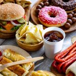 A spread of fast-food snacks: cheeseburgers, a pink-frosted donut, tortilla chips with dip, sliced bread, sausages, and other baked goods on wooden boards.