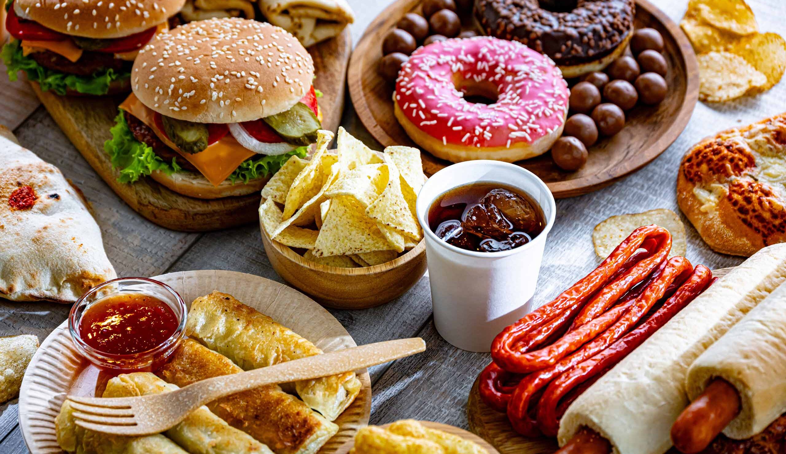 A spread of fast-food snacks: cheeseburgers, a pink-frosted donut, tortilla chips with dip, sliced bread, sausages, and other baked goods on wooden boards.