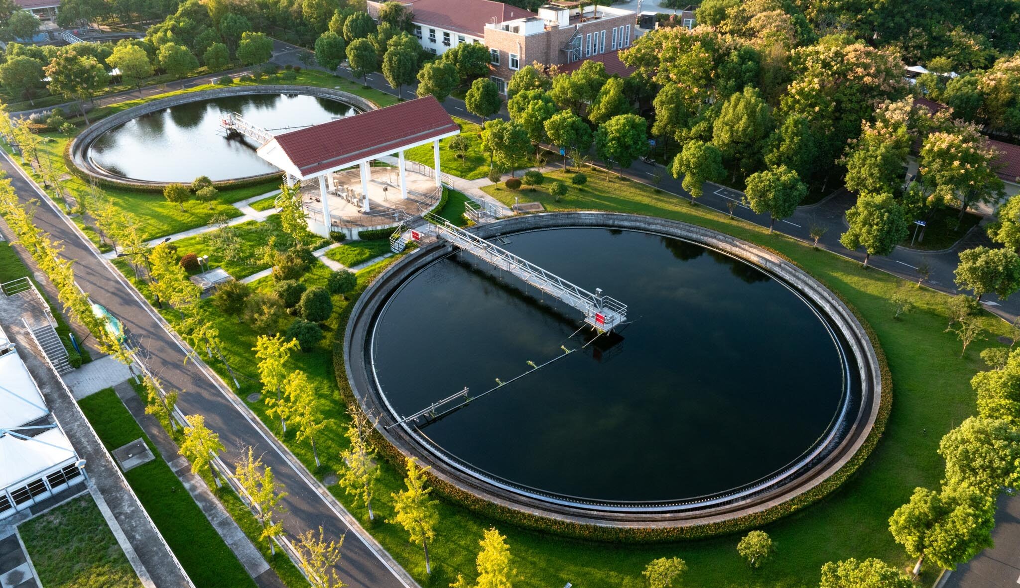 Aerial view of a circular wastewater treatment tank with a red-roof pavilion and surrounding green park area.