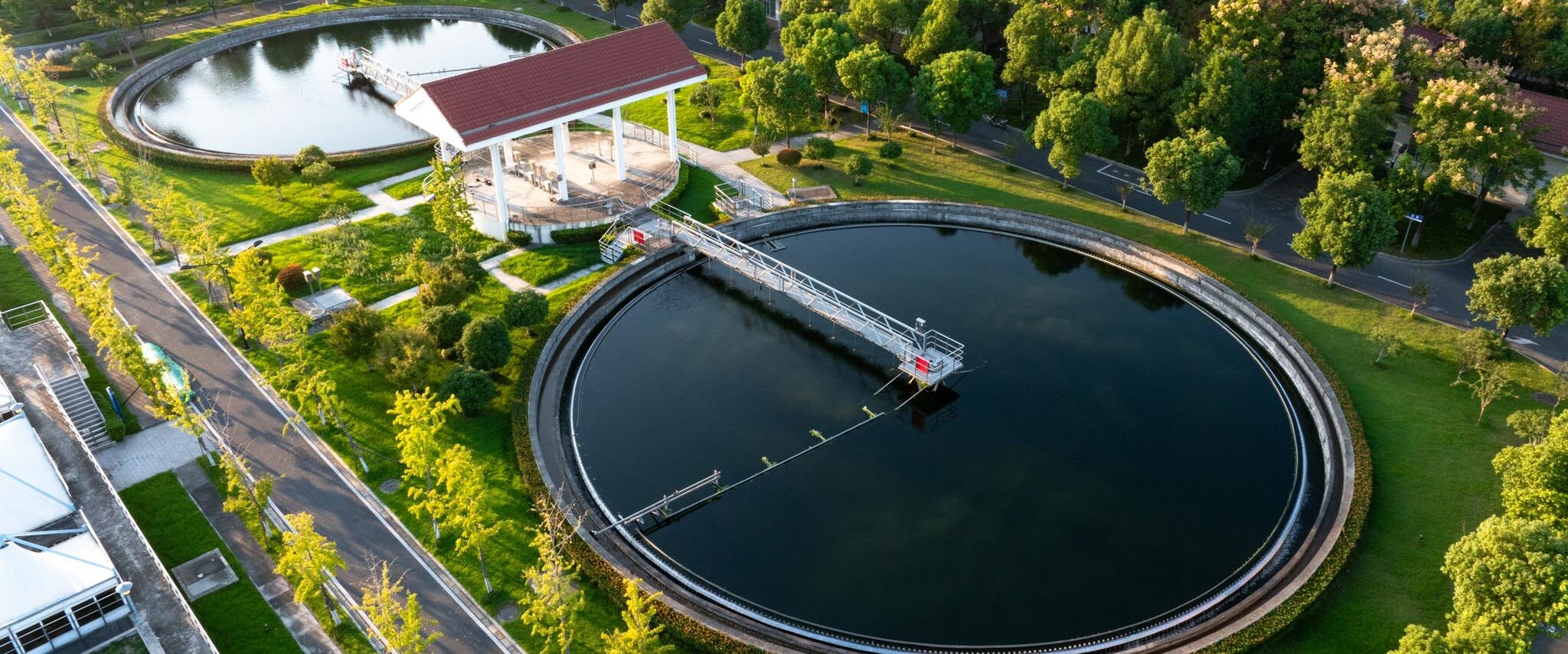 Strategic implications for wastewater OEMS. Aerial view of a wastewater treatment facility with two large circular sedimentation tanks, a central raised walkway, and a red-roofed shelter, surrounded by green lawns and tree-lined roads the facility.
